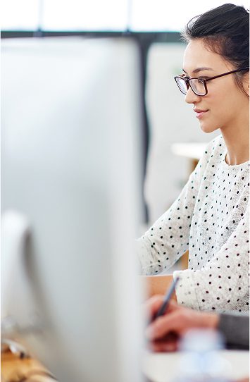 A woman with glasses and a white, polka-dotted sweater works at a computer, focused on web design development. The image is bright and modern, with a blurred foreground of a large monitor.