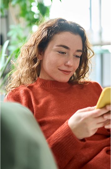 A woman with wavy brown hair, wearing an orange sweater, is sitting indoors and smiling while looking at her yellow smartphone, possibly browsing logos and branding inspiration. Green plants and natural light are visible in the background.