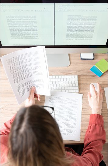 A person sits at a desk with a computer, reviewing printed documents while using a mouse. Two monitors display content marketing and copywriting materials. A keyboard, sticky notes, and a phone are also on the desk.