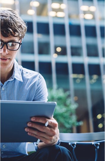 A person wearing glasses and a striped shirt reads from a tablet outdoors, possibly reviewing notes for clients, with a modern office building blurred in the background.